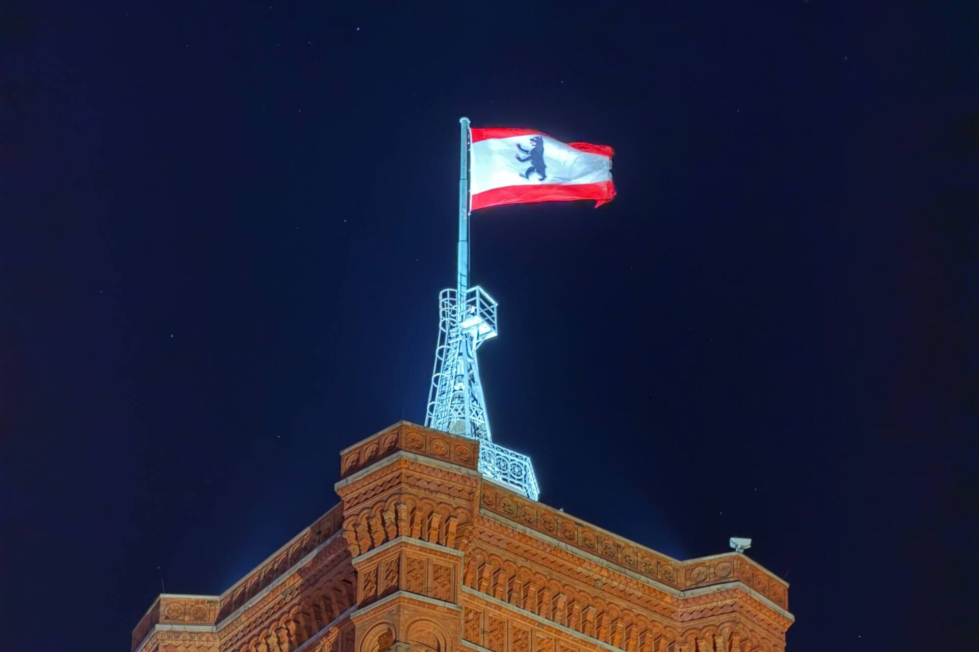 Turm eines Backsteingebäudes bei Nacht mit beleuchtetem Wappenflaggenmast und Uhrfenstern